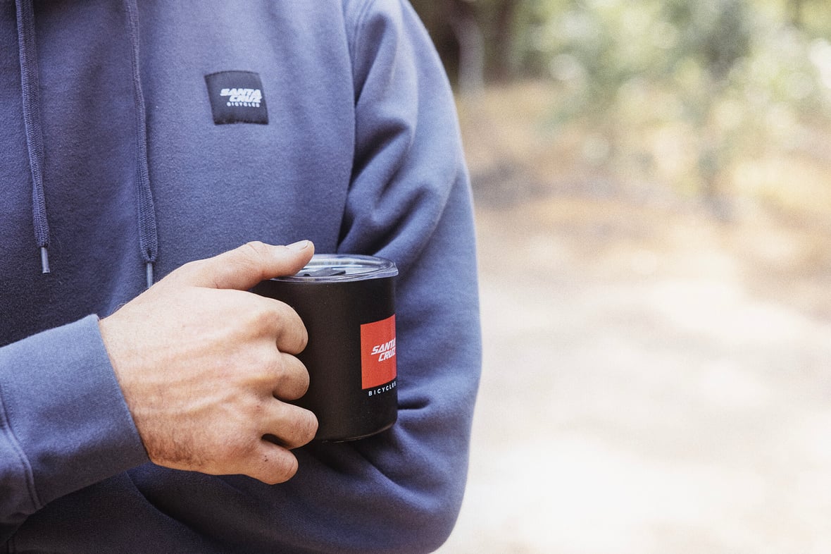 Hand models a black Cannery Cup against the backdrop of a sweet Patch Hoodie in Maritime Blue